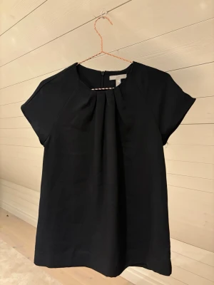 Svart topp från H&M med veckad front - En stilren svart topp med korta ärmar och snygg veckad detalj framtill. Toppen har rund halsringning och dragkedja bak. Perfekt till jeans eller kjol för en clean look.