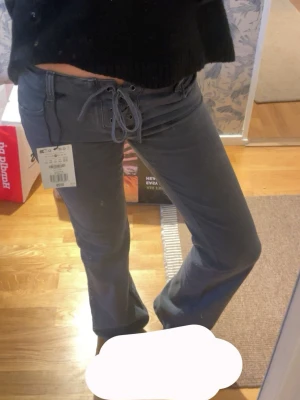 Bootcut jeans med snörning - Snygga mörkgrå jeans med bootcut passform och låg midja. Jeansen har en unik snörning framtill i midjan som ger en cool detalj. Perfekta för dig som vill ha något annorlunda i garderoben.