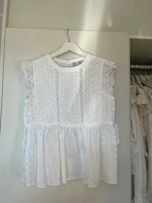 Vit broderad blus med spetsdetaljer - Supersöt vit blus med broderade detaljer och volangkant. Ärmlös modell med spets längs axlarna och knyt i midjan ❣️