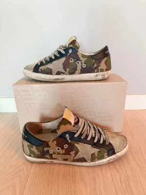 Golden Goose sneakers med camouflage - Golden Goose sneakers med coolt camouflage-mönster i grönt, brunt och beige. Skorna har en stjärndetalj på sidan, svarta skinnpartier vid hälen och snörning med logotyp på skosnörena. Platt sula och rund tå. Perfekt för dig som gillar streetstyle och unika detaljer. Skick 8/10 det ser värre ut på bilderna på moccan än va de är i verkligheten 