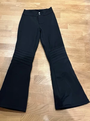 Svarta skidbyxor från 8848 Altitude - Snygga svarta skidbyxor från 8848 Altitude med utsvängda ben och ribbade detaljer över knäna. Byxorna har hög midja, två knappar framtill och är perfekta för vinterns alla äventyr och för att åka skidor. Materialet är slitstarkt och håller dig varm under kalla dagar.