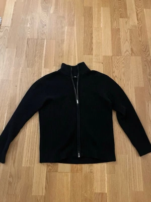 Svart full zip tröja - Svart ribbstickad tröja med dragkedja framtill och hög krage. Tröjan har långa ärmar och en rak passform, perfekt för lager-på-lager-stil. Enkel och stilren design som funkar till det mesta.