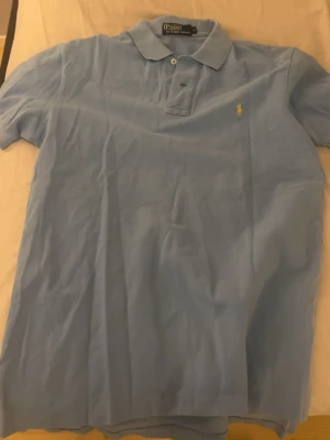 Ljusblå pikétröja från Ralph Lauren - Klassisk ljusblå pikétröja från Ralph Lauren med korta ärmar och krage. Tröjan har den ikoniska gula broderade loggan på bröstet och stängs med två knappar framtill. Tillverkad i bomull för en skön och luftig känsla.