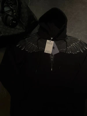 Svart hoodie Marcelo Burlon med vingar - Säljer en svart hoodie från Marcelo Burlon County of Milan med ikoniskt vingtryck över axlar och bröst. Hoodien har dragsko i huvan och är tillverkad i mjukt bomullsmaterial. Perfekt för dig som vill sticka ut med en unik design. Skickas självklart i original förpackningen!