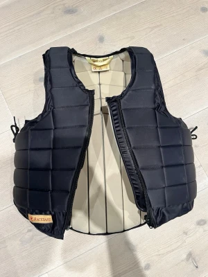 Racesafe ridväst  - Säljer en svart ridväst från Racesafe med vadderad design och dragkedja framtill. Västen har justerbara snören i sidorna och är tillverkad i slitstarkt material. Har ett litet märke på insidan av västen, annars helt felfri. Storlek: XS Orginalpris: 3 700kr 