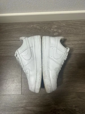 Vita Nike Air Force 1 sneakers - Klassiska vita Nike Air Force 1 sneakers med låg profil och rund tå. Skorna har snörning och är tillverkade i skinn med den ikoniska swoosh-loggan på sidan. Perfekta för en clean och stilren look.