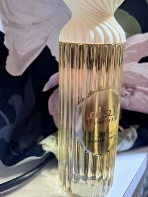 Lattafa Najdia Parfymmist - Lattafa Angham parfymmist i en elegant, räfflad parfymflaska med guldfärgad etikett och vit, skulptural kork. Flaskan har en lyxig känsla och passar perfekt för dig som gillar unika dofter och snygg design på parfymflaskor.