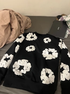 Denim tears tröja pass xs-s - Cool svart sweatshirt med stora vita blommor tryckta över hela tröjan. Rund halsringning och långa ärmar med ribbade muddar. Perfekt för dig som vill sticka ut med ett unikt mönster och streetwear-vibe.
