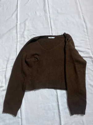 Brun stickad tröja från NA-KD - Mysig brun stickad tröja från NA-KD i croppad lite modell. Tröjan har v-ringning och långa ärmar, perfekt för lager på lager under höst och vinter. Enkel och stilren design som är lätt att matcha med jeans eller kjol, den är även ganska baggy. 