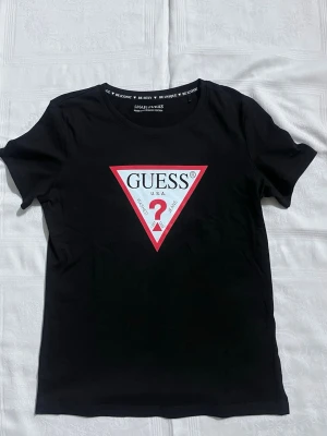 Svart GUESS t-shirt med logga - ALDRIG ANVÄND, Svart t-shirt från GUESS med den ikoniska trekantsloggan i vitt och rött framtill. Tillverkad i 100% ekologisk bomull och har rund halsringning samt korta ärmar. Perfekt för dig som gillar klassisk streetstyle med en twist.