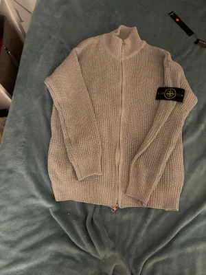 Grå stickad kofta från Stone Island - Säljer en grå stickad kofta från Stone Island med dragkedja framtill och ribbade muddar. Klassisk patch på ärmen och hög krage för extra stilpoäng. Perfekt till lager på lager under höst och vinter.