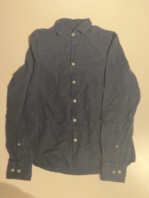 Blå skjorta från J.Lindeberg - Säljer en stilren blå skjorta från J.Lindeberg i storlek S. Skjortan har klassisk krage, vita knappar framtill och långa ärmar med knapp vid manschetten. Tillverkad i mjuk bomull som känns skön mot huden. Perfekt för dig som gillar enkel och clean stil.