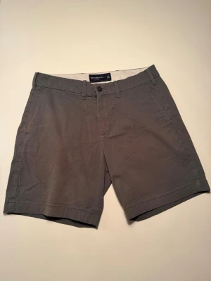 Grå shorts från Abercrombie & Fitch - Snygga grå shorts från Abercrombie & Fitch med klassisk passform. De har bälteshällor, knapp och dragkedja framtill samt två sidofickor och två bakfickor med knapp. Tillverkade i mjuk bomull, perfekta för sommardagar.