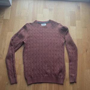 Rostbrun stickad tröja med struktur - Snygg rostbrun stickad tröja från Oscar Jacobson med diskret strukturmönster över hela plagget. Tröjan har rund halsringning, långa ärmar och ribbstickade muddar vid ärmslut och nederkant. Perfekt för lager-på-lager-stil.