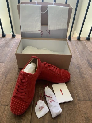 Röda sneakers från Christian Louboutin - Säljer ett par röda sneakers från Christian Louboutin i mocka med matchande röda nitar på tån. Skorna har snörning och en klassisk låg profil. Kommer med originalkartong, dammpåsar och extra tillbehör. Perfekta för dig som vill sticka ut.