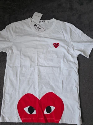 Vit Comme des Garçons Play t-shirt - Säljer en vit t-shirt från Comme des Garçons Play med det ikoniska röda hjärtat med ögon på bröstet och ett stort hjärta med ögon tryckt längst ner. Klassisk passform och rund hals, perfekt till jeans eller shorts.