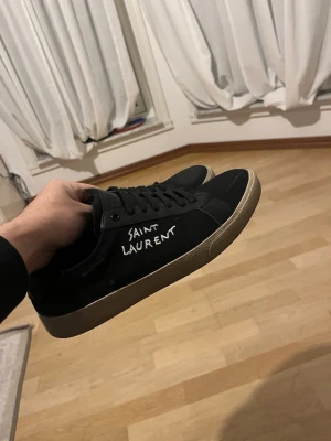 Svarta sneakers - Säljer ett par svarta sneakers med vit logga på sidan. Skorna har klassisk låg profil, rund tå och svart snörning. Yttermaterialet är troligtvis syntet och sulan är beige. Perfekta för dig som gillar stilrena och exklusiva sneakers. Lite avfärgning på ena skon ses på bild. Men är inte så synligt, därför säljs billigt. Obs endast skorna.