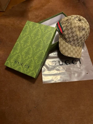 Ingen väst – Gucci keps med GG-mönster - Det här är en beige Gucci keps med klassiskt GG-mönster och grönt/rött band på sidan. Kepsen är tillverkad i bomull och polyester och har justerbart spänne bak. Säljes med originalkartong och dustbag.