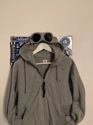 Grå hoodie med goggles från C.P. Company - Säljer en grå hoodie från C.P. Company med dragkedja framtill och två fickor. Huvan har inbyggda goggles och justerbara snören. Perfekt för dig som vill sticka ut med en unik streetwear-look.