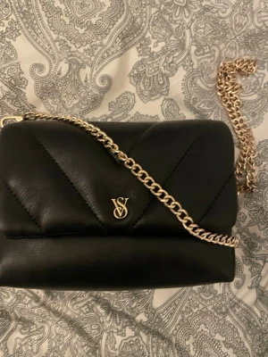 Svart axelväska från Victoria's Secret - Snygg svart axelväska från Victoria's Secret med quiltad design och guldfärgad kedja. Väskan har VS-logga i guld framtill och är tillverkad i skinnimitation. Perfekt för att lyfta din outfit med en trendig touch.