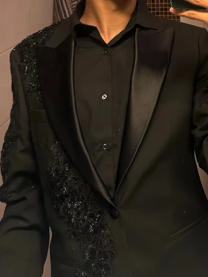 Svart kavaj med glittriga detaljer - ASOS - Säljer en svart kavaj med breda slag och glittriga, broderade detaljer över axel och ärm. Perfekt för dig som vill sticka ut på festen. Kostymen är ifrån ASOS men har sedan sytt på detaljerna.                         Använt den en gång. Storleken på kavajen är EU 46 regular och kostymbyxorna är i storlek W30 regular