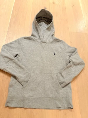 Grå hoodie från Polo Ralph Lauren - Snygg grå hoodie från Polo Ralph Lauren med klassisk huva och känguruficka framtill. Liten mörkblå logga broderad på bröstet. Tillverkad i mjuk bomullsmix som känns skön mot huden. Perfekt för en avslappnad och stilren look.     Tveka inte att höra av dig vid minsta fundering😊