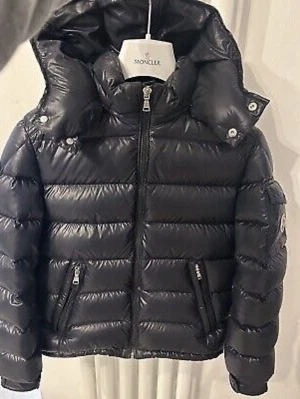 Svart dunjacka från Moncler - Snygg svart dunjacka från Moncler med glansig finish och quiltad design. Jackan har huva, hög krage och dragkedja framtill samt två fickor med dragkedja. Perfekt för kalla dagar och ger en trendig look.