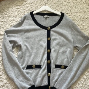 Grå kofta med svarta detaljer och guldknappar - Säljer en snygg och stilren grå kofta med svarta detaljer och guldknappar. Koftan har långa ärmar och två små fickor framtill. Perfekt för både vardag och fest! 🖤✨