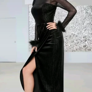 Svart glittrig långklänning med slits - Supersnygg svart långklänning med glittrigt tyg, transparenta långärmar och fluffiga detaljer vid ärmsluten. Klänningen har en hög slits på sidan och en figurnära passform som ger en elegant siluett. Perfekt för dig som vill sticka ut med stil.