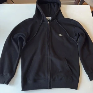 Svart hoodie från Lacoste med dragkedja - Svart hoodie från Lacoste i regular fit med klassisk krokodillogga på bröstet. Tröjan har huva med snörning, hel dragkedja och två stora fickor framtill. Perfekt för en chill och stilren look.
