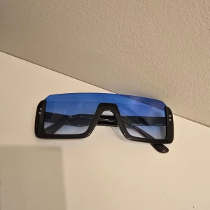Svarta solglasögon med blått visir - Coola solglasögon med svart, kantig båge och stora blåtonade visirglas. Riktigt futuristisk vibe och perfekt accessoar för att sticka ut. Snygga detaljer med små silvriga prickar på sidorna.