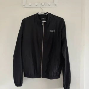 Svart vindjacka från BLACC - Svart vindjacka från BLACC med stilren design och logga på bröstet. Jackan har hel dragkedja framtill, elastiska muddar och är tillverkad i lätt polyester. Perfekt för dig som gillar enkel och sportig stil.
