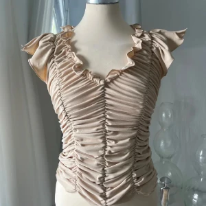 Beige omlott topp med volang - Säljer en beige omlott topp med snygga volangdetaljer vid axlar och halsringning. Toppen har ett rynkat tyg framtill som ger en cool och trendig look. Materialet är mjukt och stretchigt, perfekt för dig som gillar att sticka ut med din stil.