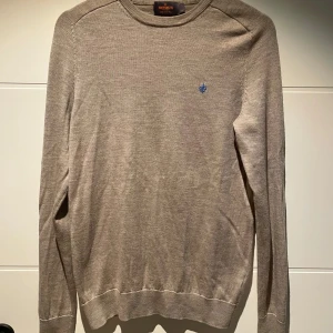 Beige tröja i merinoull från Morris - Säljer en stilren beige tröja från Morris i 100% merinoull. Tröjan har en liten blå broderad detalj på bröstet och långa ärmar. Perfekt för kyligare dagar. 🧥