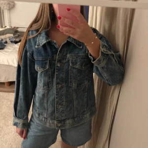 Blå jeansjacka med knappar - Säljer en klassisk blå jeansjacka med snygga metallknappar framtill och två bröstfickor. Jackan har lång ärm och en cool rynkad detalj i ryggen som ger extra stilpoäng. Perfekt att slänga över en hoodie eller t-shirt för en chill look.