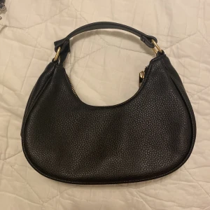 Svart axelväska från Bik Bok - Pettite svart väska från Bik Bok med rundad form och guldfärgade detaljer. Väskan är tillverkad i skinnimitation med struktur och har en praktisk dragkedja upptill. Verkligen suuuper gullig!😍😍