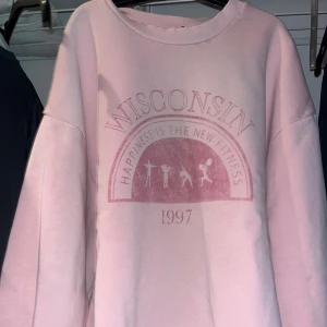 Rosa sweatshirt med Wisconsin-tryck - Säljer en oversized ljusrosa sweatshirt med trycket 'Wisconsin' och texten 'Happiness is the new fitness' samt årtalet 1997 på framsidan. Tröjan har rund hals och långa ärmar, perfekt för chill dagar. Mjuk bomullskänsla och relaxed passform.