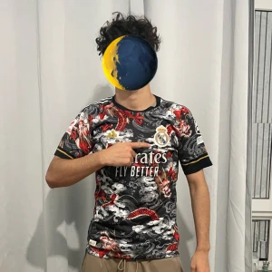 Real Madrid Ronaldo 7 svart draktröja - Säljer en unik svart Real Madrid fotbollströja med Ronaldo 7 på ryggen. Tröjan har ett fett drakmönster i rött och vitt, samt gulddetaljer på ärmarna. Materialet är lätt och andas, perfekt för match eller träning. Officiell klubbmärke på bröstet och snygg passform.