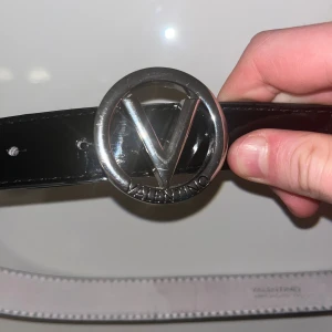 Valentino bälte - Snyggt svart bälte från Valentino med en blank finish och ett runt silverfärgat spänne med ikonisk V-logga. Bältet är smalt och tillverkat i skinn, perfekt för att lyfta din outfit med en lyxig touch. Passar till jeans, kjol eller klänning för en trendig look.