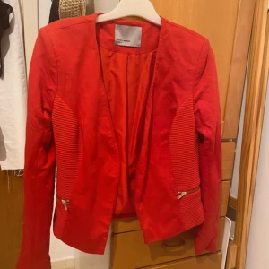 Röd kavaj från Vero Moda - Snygg röd kavaj från Vero Moda med ribbade detaljer på sidorna och guldfärgade dragkedjor vid fickorna. 