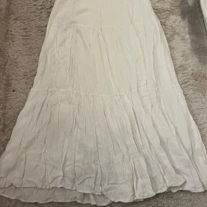 Vit långkjol i bomull - Säljer en vit långkjol med mjuka volanger och lätt rynkad struktur. Kjolen är tillverkad i tunn bomull som ger ett luftigt och flowy intryck. Perfekt för dig som gillar en enkel och clean look med lite boho-vibe.