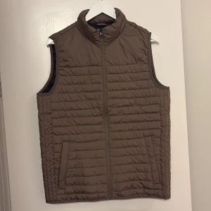 Brun dunväst från Jack & Jones M - Snygg brun dunväst från Jack & Jones i storlek M. Västen har quiltad design, hög krage och dragkedja framtill. Perfekt att slänga över en hoodie för extra värme. Två fickor på sidorna och stilren look.