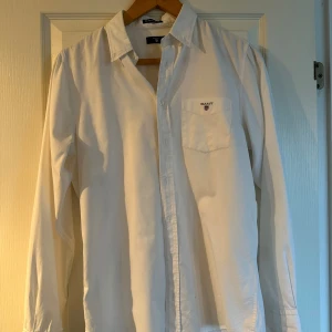 Vit långärmad skjorta från GANT - Vit klassisk skjorta från GANT, modell The Archive Oxford. Skjortan har lång ärm, bröstficka med broderad logga och knappar framtill. Tillverkad i mjuk bomull med stilren krage och enkel design som funkar till många olika looks.