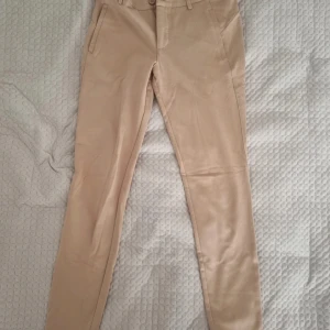 Beiga chinos med slim fit - Snygga beiga chinos med smal passform och klassiska fickor fram och bak. Byxorna har bälteshällor och stängs med knapp och dragkedja. Perfekta för en clean och stilren look. Materialet känns mjukt och bekvämt.