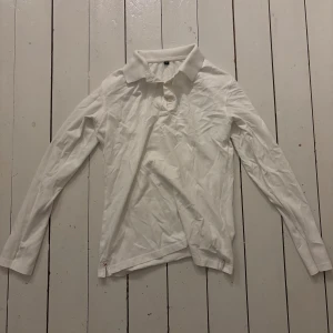 Vit långärmad pikétröja - Klassisk vit långärmad pikétröja med krage och knäppning framtill. Tröjan är i mjuk bomull och har en stilren look som passar perfekt till jeans eller chinos. Enkel och tidlös design som funkar året runt.