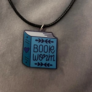 Book Worm Halsband Book Lover  - Perfekt halsband för bokälskaren! Berlock i akryl av en blå bok med texten ”book worm”. Berlocken är ca 2,5 x 2,8 cm. Trädd på ett svart snöre justerbart 45-50 cm. 