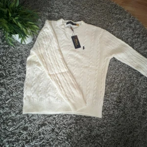 Vit kabelstickad tröja Polo Ralph Lauren - Snygg vit kabelstickad tröja från Polo Ralph Lauren med klassisk rund hals och broderad logga på bröstet. Tröjan har lång ärm och ribbade muddar. Perfekt för dig som gillar stilrena och tidlösa plagg med premiumkänsla.