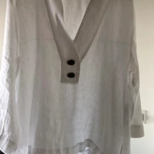 Vit linnehenley med knappar - Vit henley-skjorta i linne med lång ärm och två svarta knappar framtill. Skjortan har en djup v-ringning och är lätt transparent med rak passform. Perfekt för dig som gillar stilrena och luftiga plagg med en modern touch.