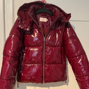 Vinröd pufferjacka från Nelly XXS - Snygg vinröd pufferjacka från Nelly i glansigt material. Jackan har huva med snörning, dragkedja framtill och tryckknappar vid fickorna. Perfekt för kalla dagar och har en cool, oversized look som sticker ut.