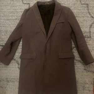 Brun kappa från J.Lindeberg - Snygg brun kappa från J.Lindeberg med klassisk krage och två framfickor. Insidan är quiltad för extra värme och komfort. Perfekt för dig som vill ha en stilren och tidlös look under höst och vinter.